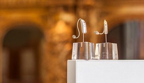 Zwei moderne Hörgeräte stehen auf transparenten Ständern vor unscharfem Hintergrund.